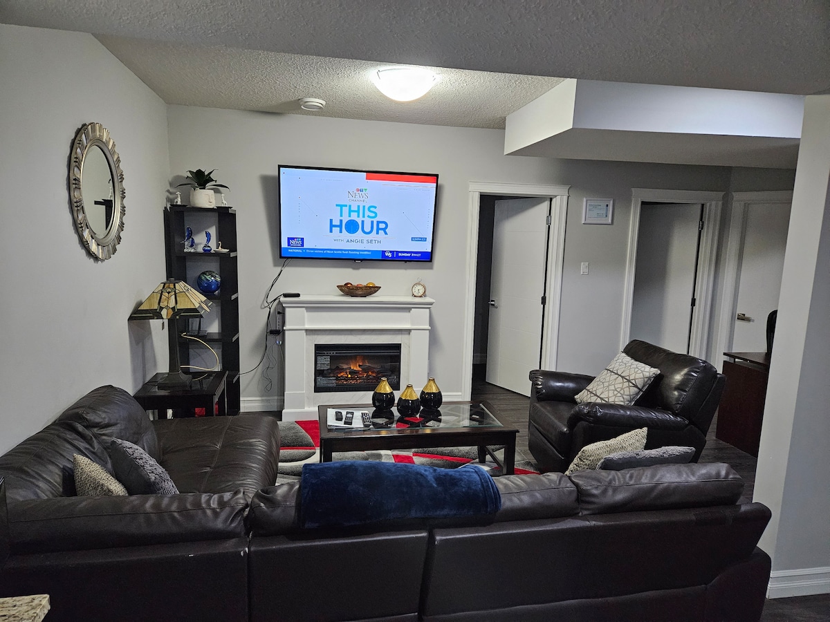 A comfortable living area is presented with dark leather couches arranged around a coffee table. A mounted flat-screen TV displays a news program above an electric fireplace. Decorative items and a lamp enhance the corner, while soft lighting creates a welcoming ambiance.