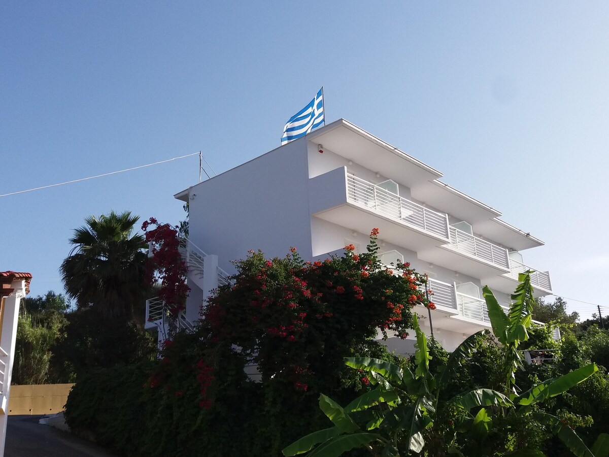 A three-story white building is surrounded by lush greenery and vibrant flowering plants. A Greek flag is prominently displayed on the top floor, and balconies can be seen on each level, providing open spaces that frame the tranquil setting.