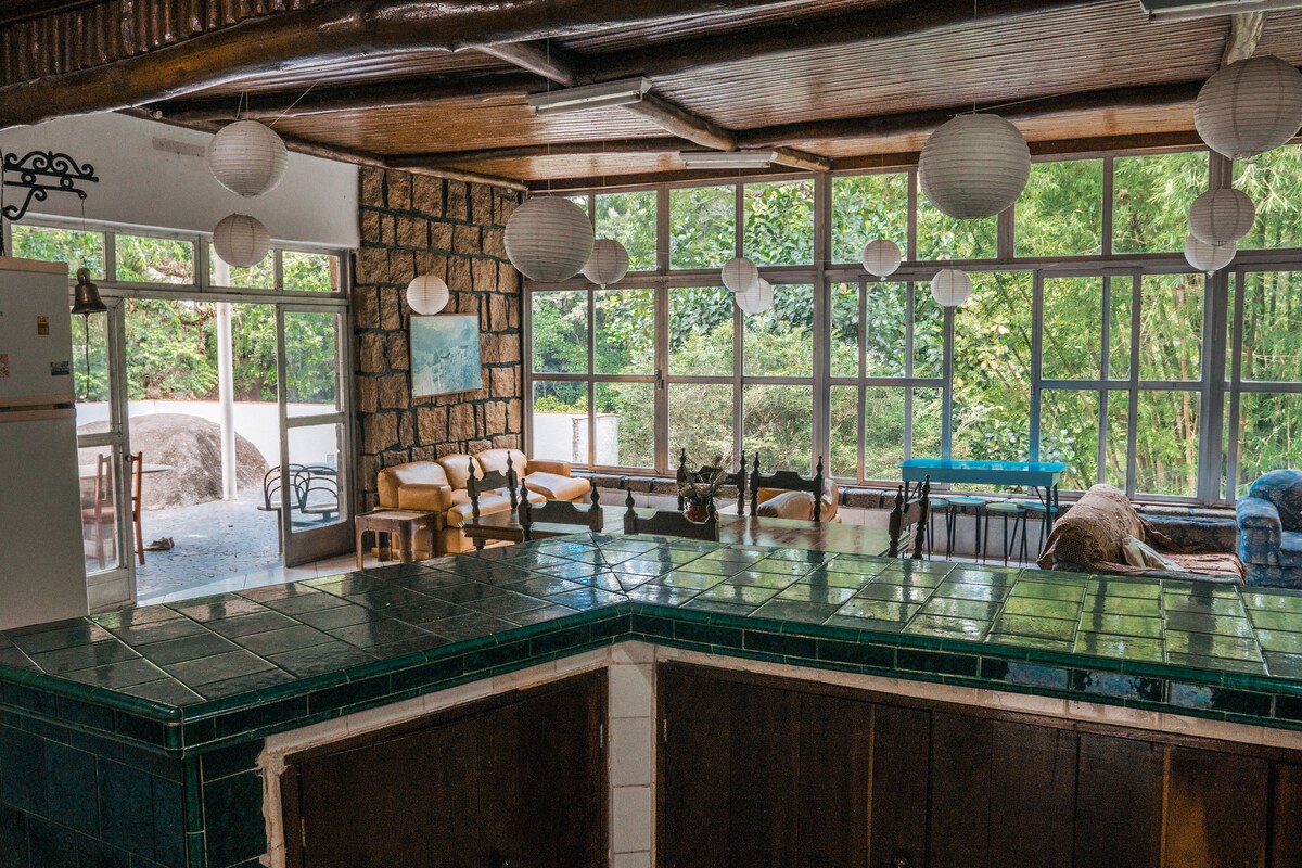 A spacious living area is illuminated by natural light filtering through large windows, with views of the greenery outside. The area features a cozy seating arrangement and a dining table, while pendant lights add a modern touch. A tiled counter provides a functional kitchen space.