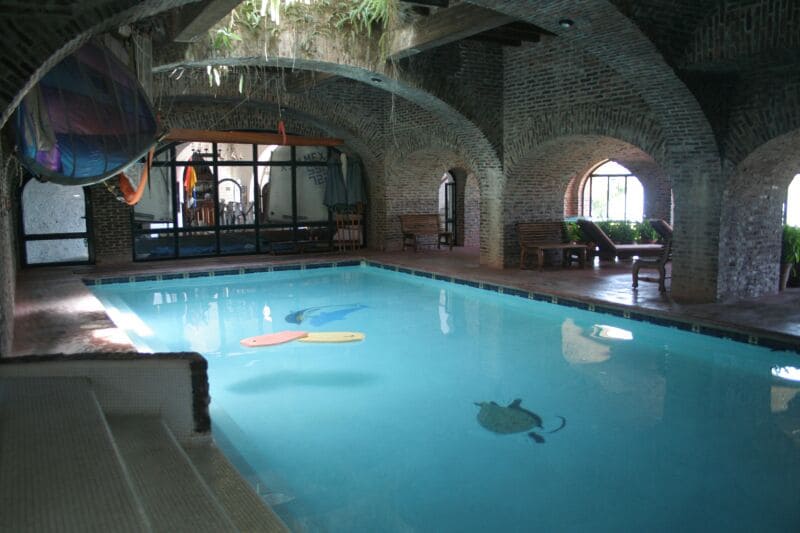 An indoor swimming pool is framed by rustic brick arches and large windows. Colorful surfboards float on the surface, with light reflecting off the clear water. Wooden benches are placed along the sides, offering a comfortable space to relax and enjoy the serene atmosphere.