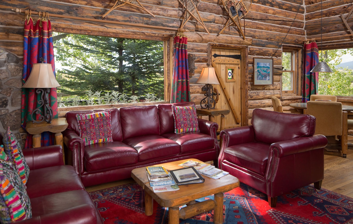 A warm living area is presented with rich red leather sofas and vibrant patterned cushions. A wooden coffee table sits in the center, surrounded by a colorful area rug. Large windows frame views of the lush greenery outside, enhancing the cabin's natural ambiance.