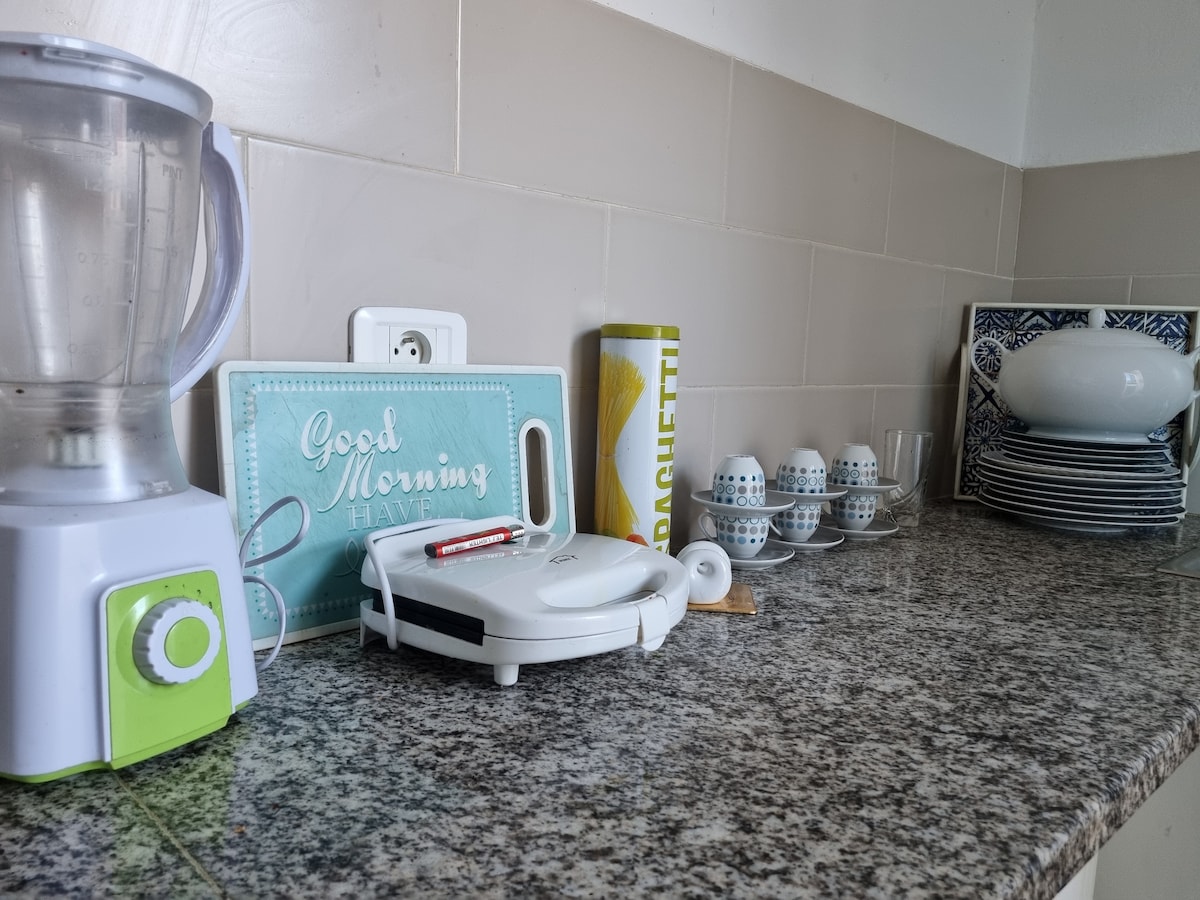 A well-organized kitchen counter displays a blender, a sandwich maker, and a canister labeled 'Spaghetti.' A cheerful sign reading 'Good Morning' is visible. Several plates and cups are neatly arranged, complemented by a serving dish, highlighting the apartment's functionality for meal preparation.