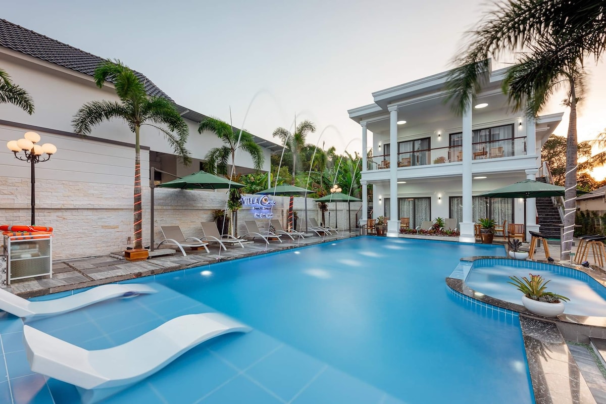 A spacious pool area is framed by palm trees and lounge chairs. A modern building rises in the background with multiple balconies overlooking the water. Water features create gentle fountains, enhancing the serene environment of the pool deck.