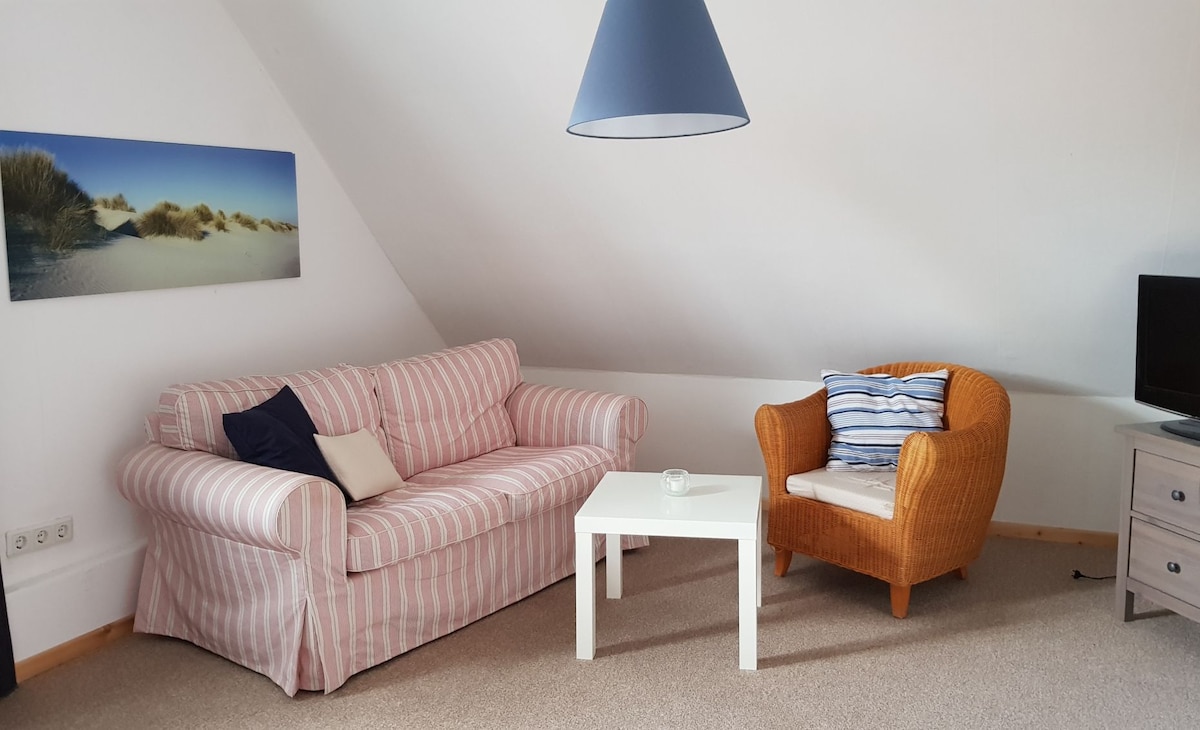 A cozy living area is featured, displaying a striped sofa and a textured armchair. A simple white table is positioned between them. Soft natural light filters in from above, enhancing the serene setting. A flat-screen TV and a wall art piece showcasing a beach scene are present.
