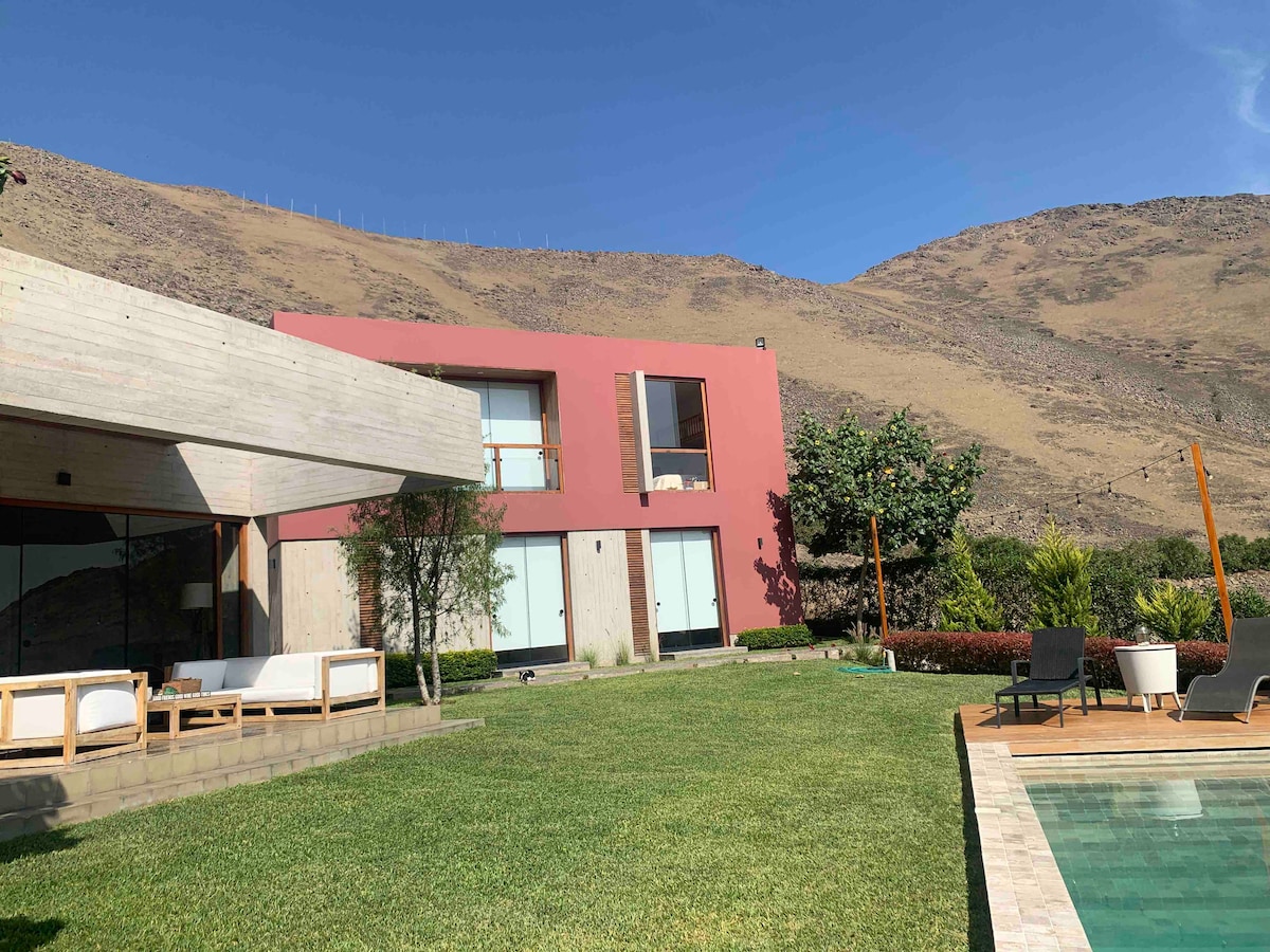 The modern structure features a combination of concrete and vibrant red exterior, set against a backdrop of rolling hills. The spacious lawn is bordered by neatly maintained plants, and a swimming pool reflects the clear blue sky. Large windows provide ample natural light.