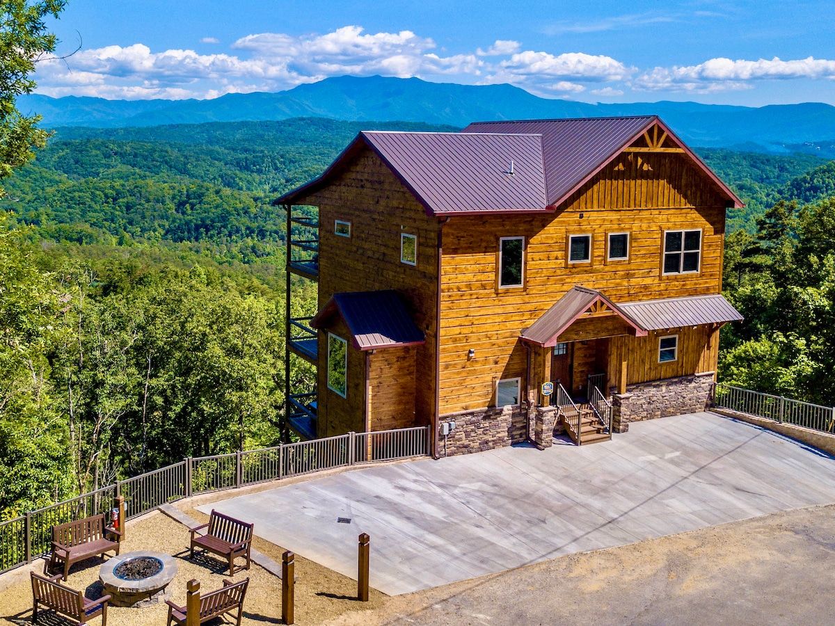 The cabin's exterior showcases a rustic wooden design, complemented by a sloped metal roof. Expansive views of the Smoky Mountains are visible in the background, while a spacious concrete area features seating and a fire pit, enhancing the outdoor experience.