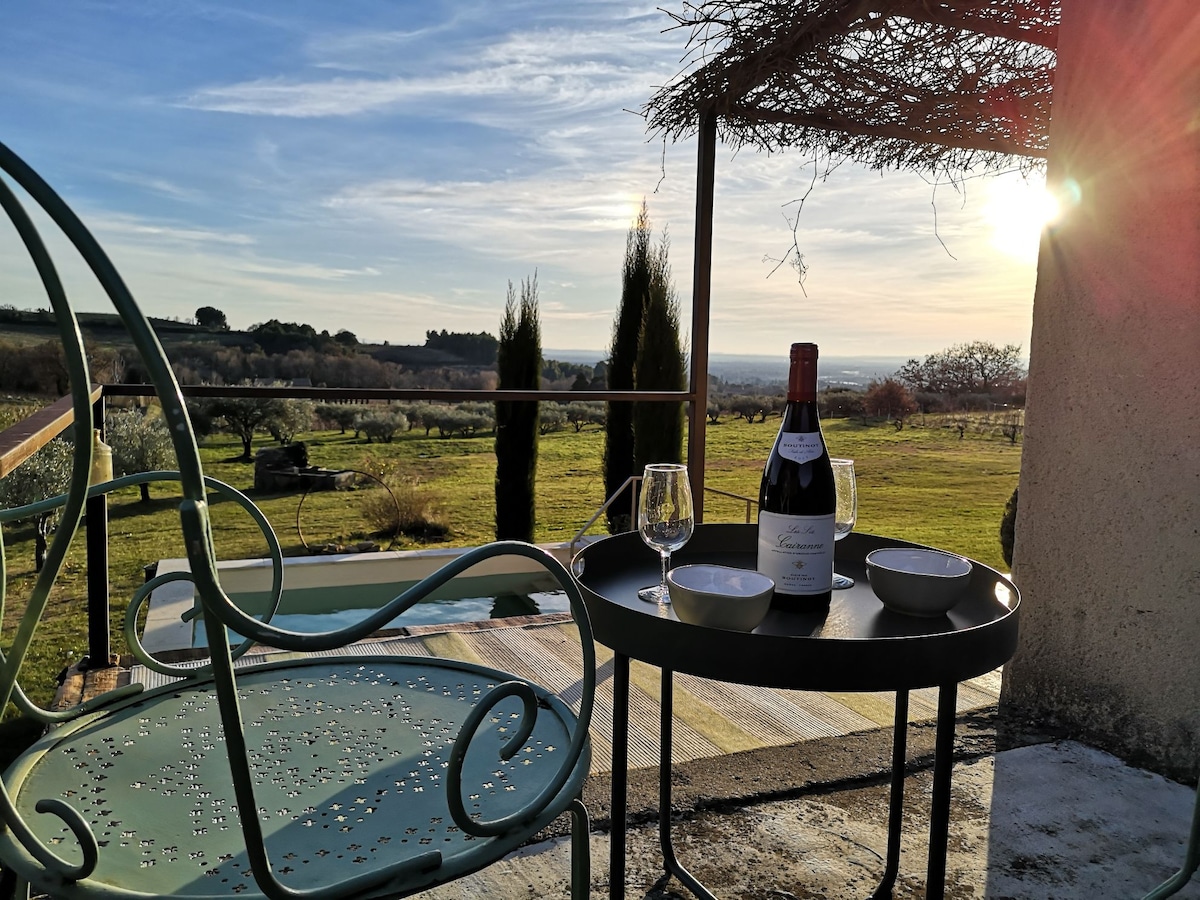 A private terrace is displayed, featuring a small round table set with a bottle of wine and two glasses. A scenic landscape of rolling hills and trees stretches into the background, with sunlight casting a warm glow over the peaceful setting.