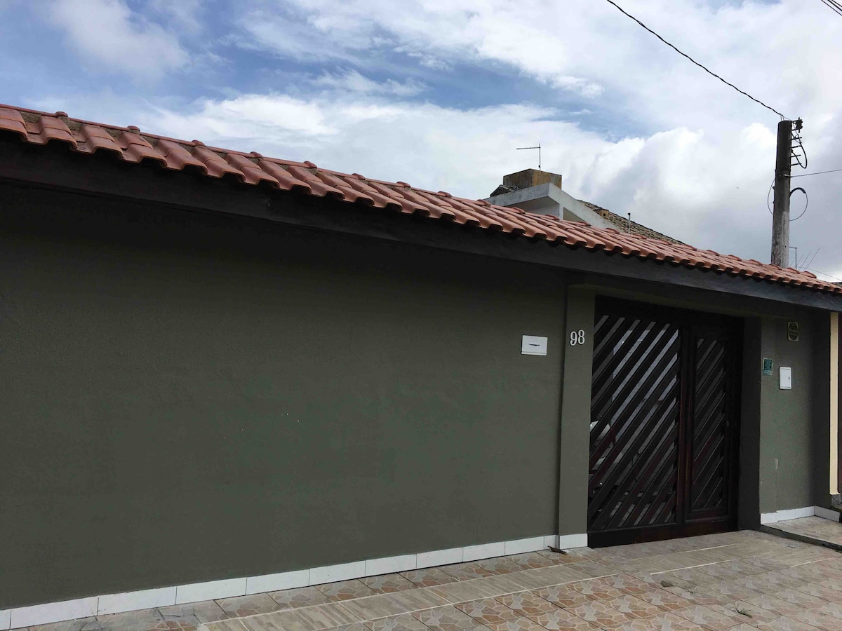 The exterior of the house is shown, featuring a neutral-colored facade with a tiled roof. A wooden front door with diagonal slats is highlighted, accompanied by a stone pathway leading to the entrance. The surrounding area includes a clear sky and hints of nearby structures.