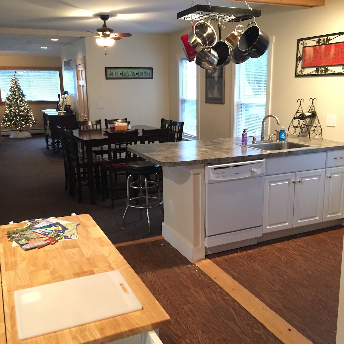 The kitchen area features a central island with a wooden countertop, complemented by a set of hanging pots above. A dining space with wooden tables and chairs is visible in the background, and a decorated Christmas tree is positioned near a large window.