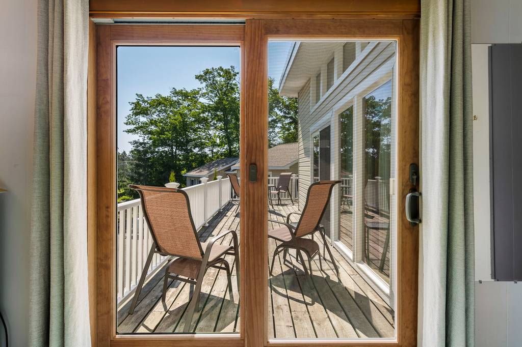A spacious deck is visible through large glass sliding doors, featuring several outdoor chairs arranged on the wooden flooring. Surrounding trees provide a backdrop, enhancing the outdoor space's natural feel.