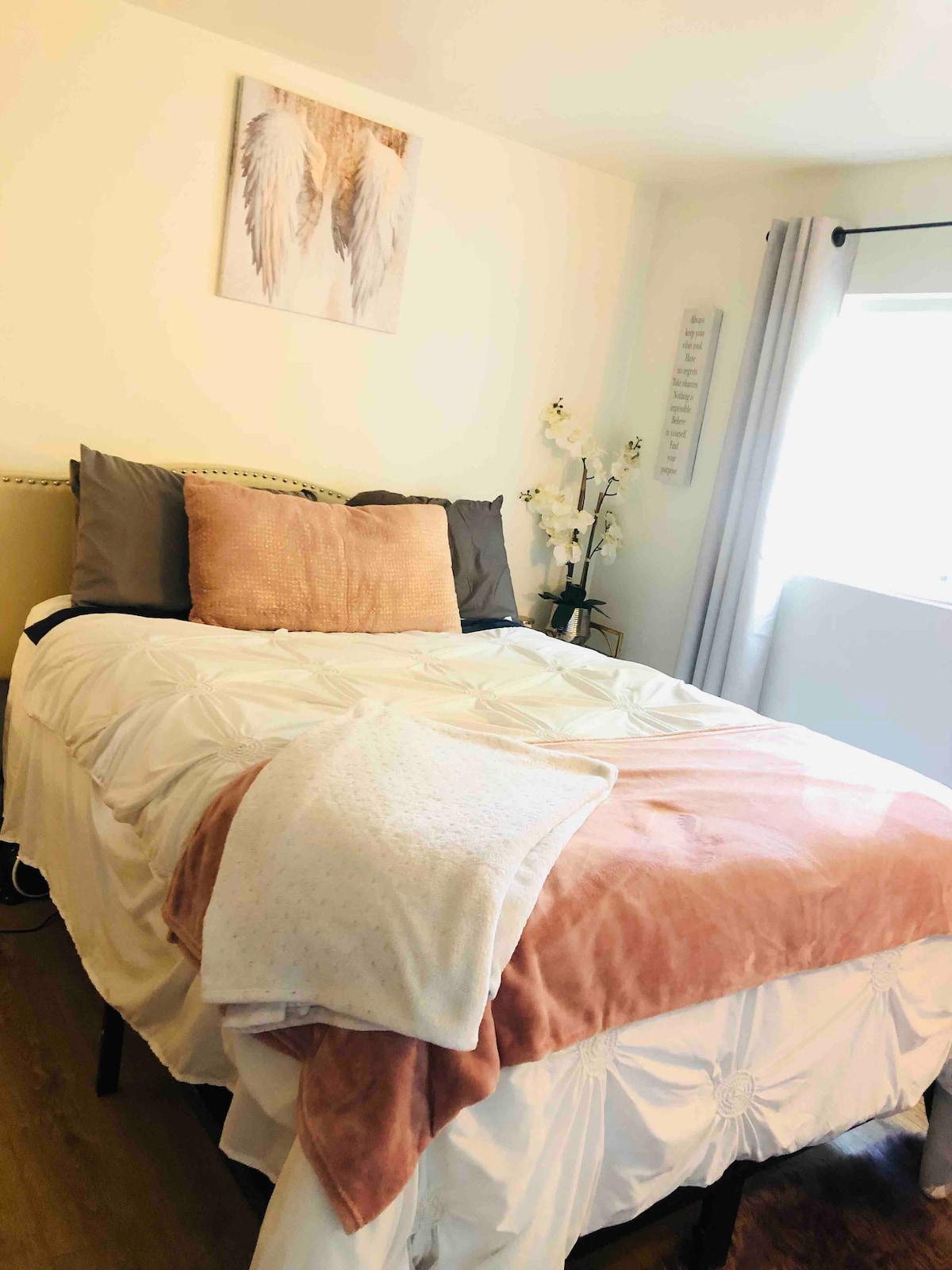 A cozy sleeping area features a spacious bed dressed in light bedding with soft accent pillows. A piece of wall art hangs above the bed, while a window allows natural light to enter, complemented by soft grey curtains. Floral decor adds a touch of warmth.