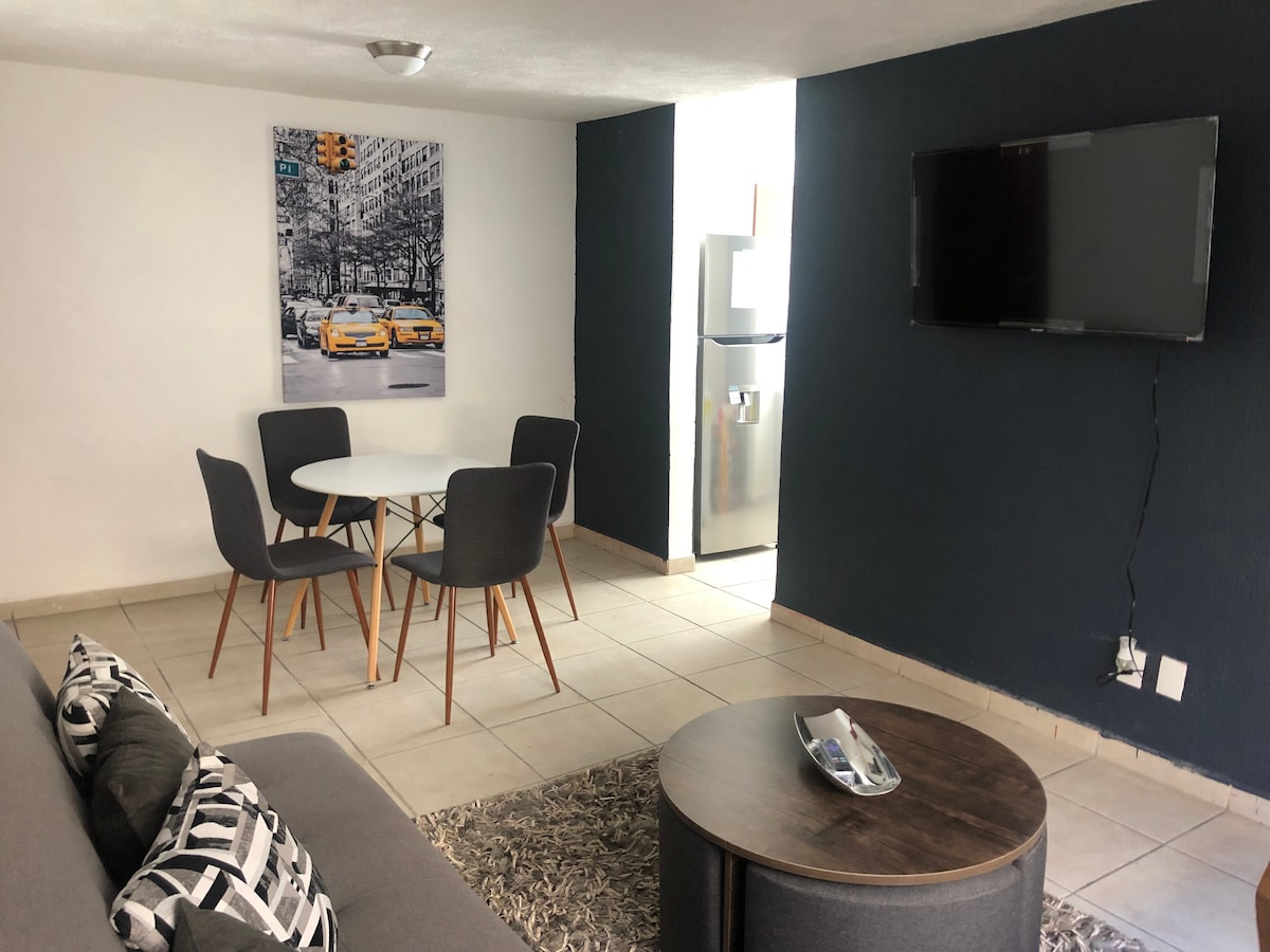 A modern living area features a circular dining table surrounded by four gray chairs. A large television is mounted on the wall opposite a spacious gray sofa with decorative cushions. A small round coffee table sits on a textured rug, with a light streaming from a nearby window.