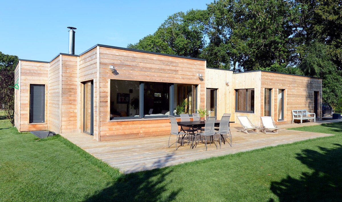 A contemporary wooden house is set amidst a grassy area, featuring large windows and a spacious wooden deck. Outdoor seating arrangements, including lounge chairs and a dining table, are positioned for relaxation. The building is surrounded by greenery, providing a serene atmosphere.