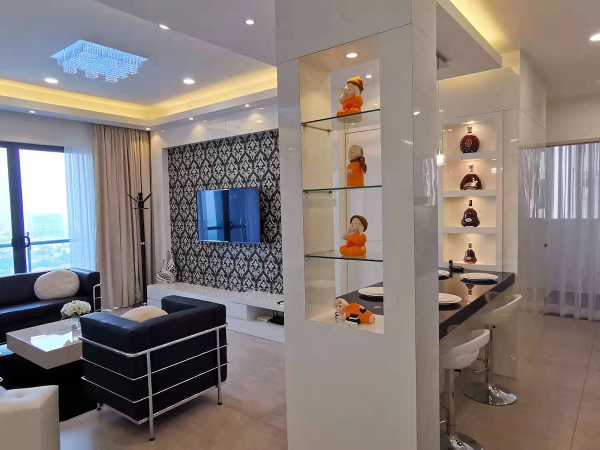 A modern living area features a comfortable seating arrangement with plush sofas and a sleek coffee table. A wall-mounted television is positioned for easy viewing. Decorative shelves showcase various ornaments, while large windows allow natural light, providing a vibrant ambiance.