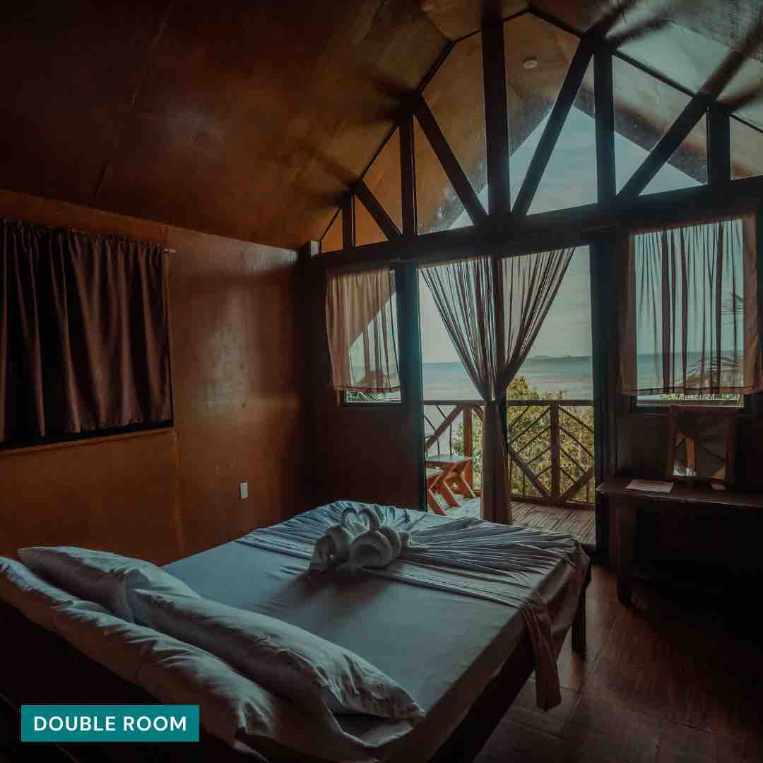 A double room is shown, featuring a spacious bed with neatly arranged linens. Large windows allow natural light to fill the space, offering views of the surrounding landscape. Wooden walls and ceiling create a warm atmosphere, complemented by curtains that frame the scenic view.