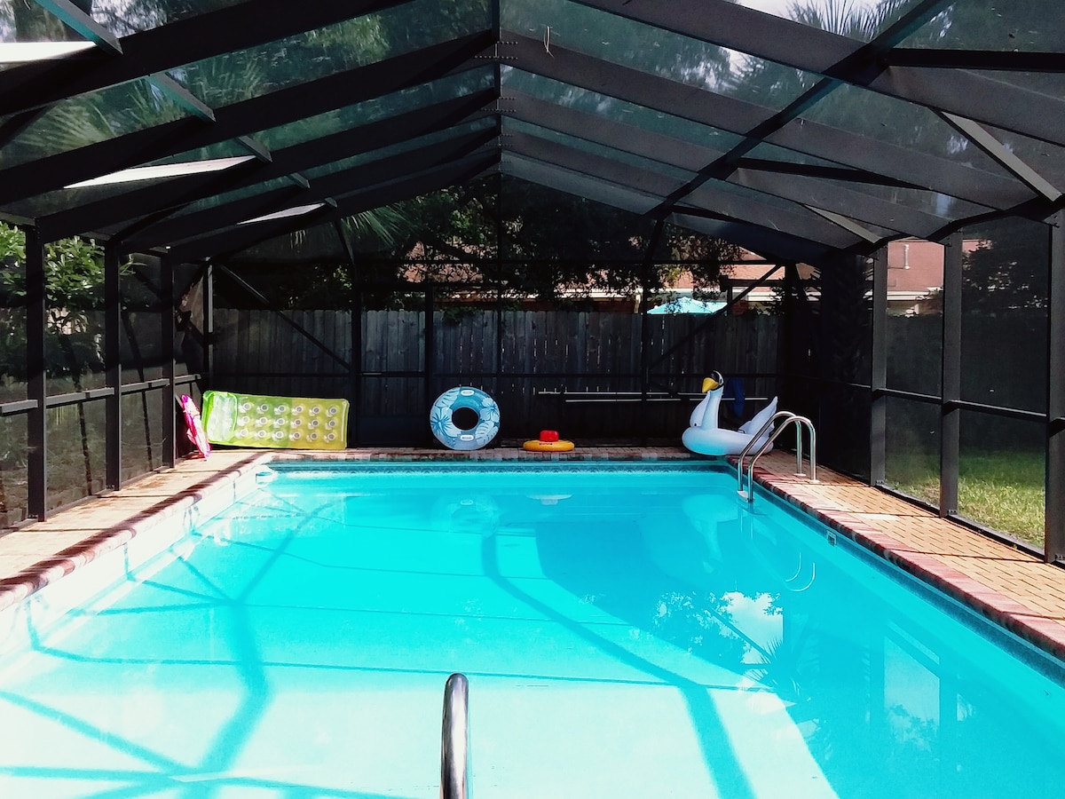 A private pool is enclosed by a screened-in structure, allowing for a comfortable swimming experience. Colorful pool floats and lounge items are placed nearby, with a bright blue water surface reflecting light. Lush greenery is visible beyond the pool area, offering a sense of privacy.