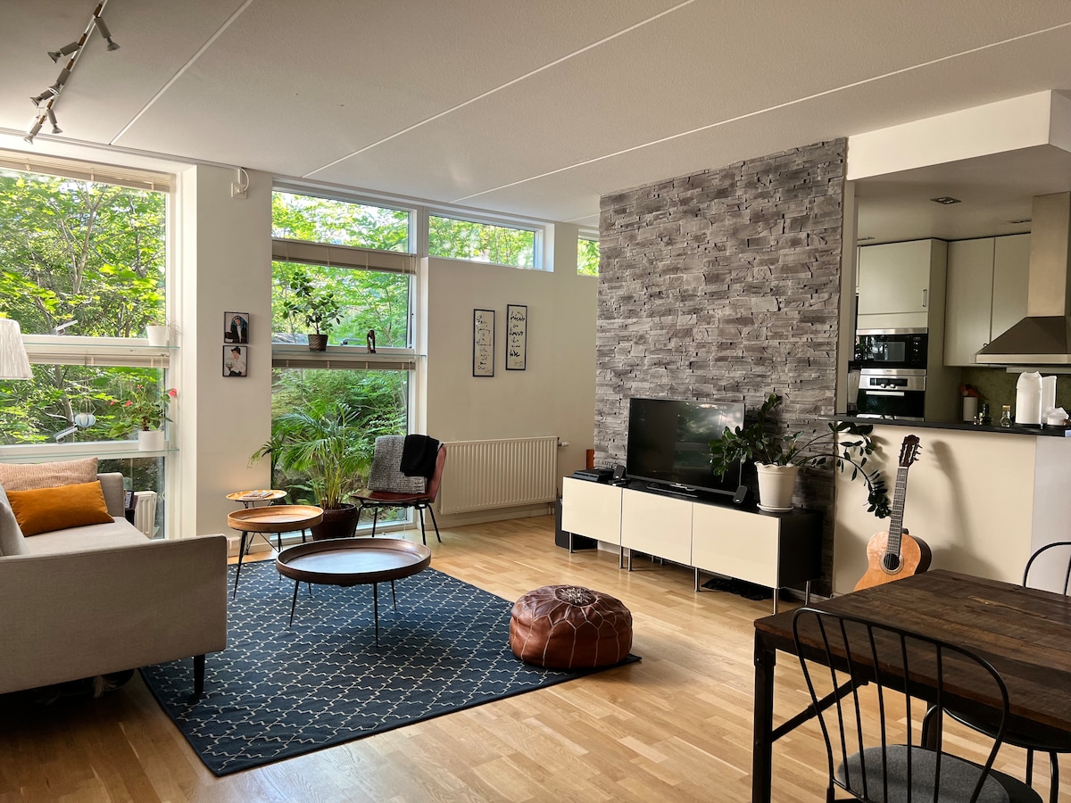 A light-filled living area features large windows overlooking greenery. A comfortable sofa is paired with a patterned rug, low wooden tables, and a modern TV unit. A guitar leans against the wall, while a dining table with black chairs occupies the adjacent space.