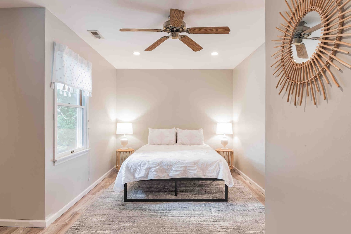 A calming bedroom features a spacious bed centered against a light-colored wall. Soft bedside lamps provide gentle illumination. Natural light filters through a window adorned with a patterned curtain. A decorative mirror adds a touch of style to the room's peaceful ambiance.