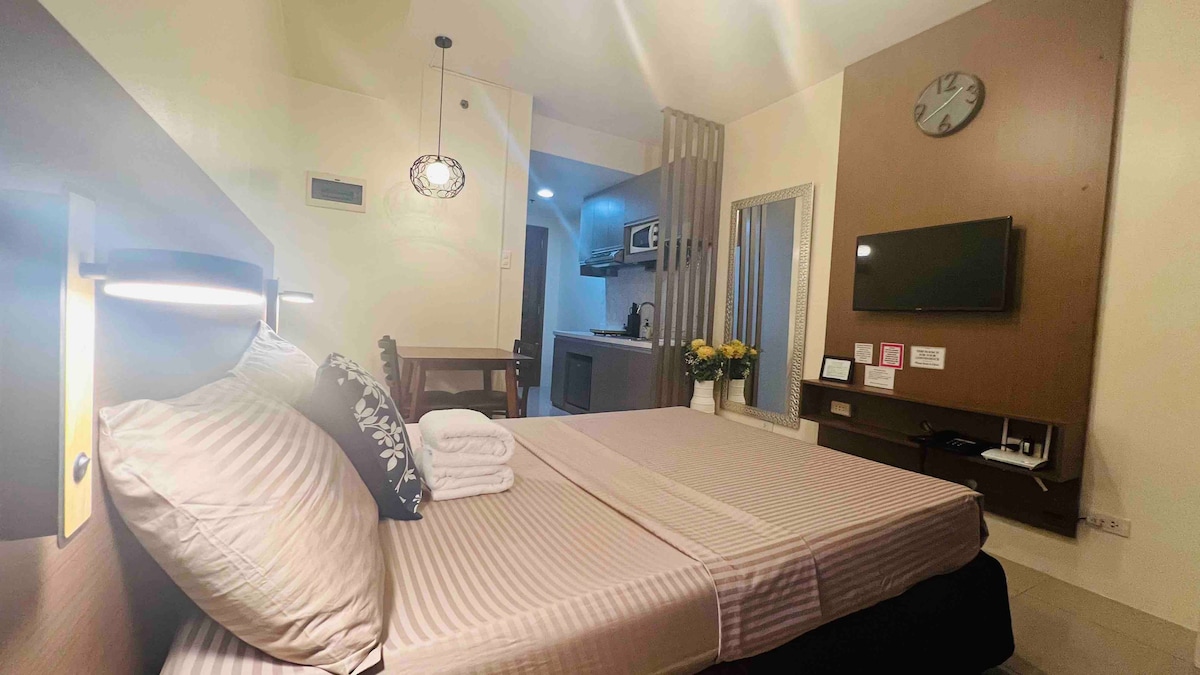 The studio unit features a spacious bed with neatly arranged towels on top. A small table and chairs are visible, complementing the kitchen area equipped with appliances. A wall-mounted TV is positioned across from the bed, and a clock adorns the wall, enhancing the cozy ambiance.