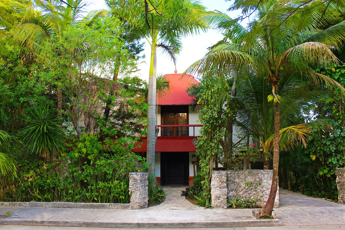 A tropical setting features a two-story building surrounded by lush palm trees and greenery. The entrance is marked by stone pillars on either side, leading to a pathway that invites guests into the property. A red-tiled roof adds a distinct architectural element.