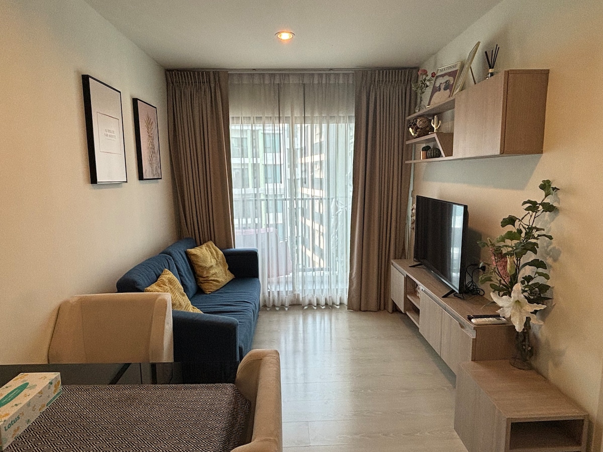 A cozy living area is furnished with a blue sofa and a beige armchair. Natural light filters through floor-to-ceiling curtains, illuminating the space. A television rests on a wooden console, complemented by decorative plants and wall art.