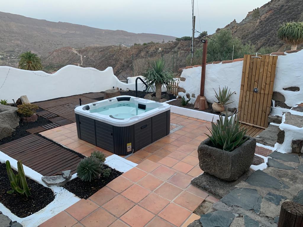 A private outdoor area features a hot tub surrounded by a terraced deck and stone pathways. Planters with various plants are positioned around the space, and the backdrop showcases a mountainous landscape under a soft sky.