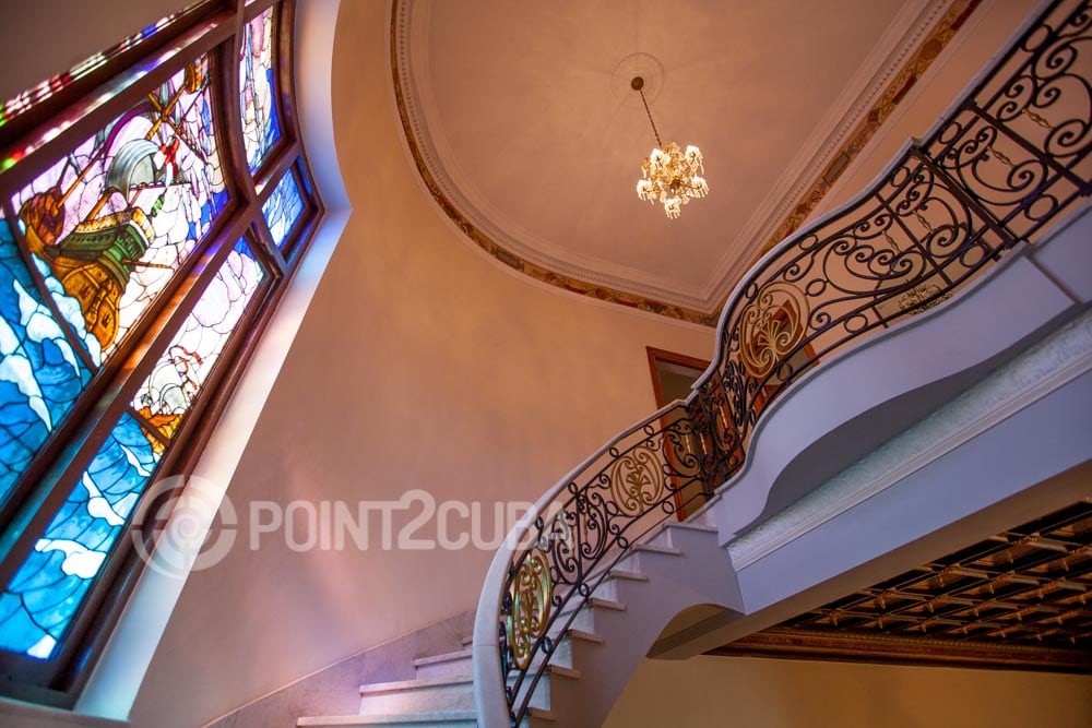 A grand staircase with an elegant design leads to an upper floor, surrounded by a vibrant stained-glass window that showcases intricate scenes. A decorative chandelier adds a touch of sophistication to the space, with warm lighting enhancing the marble flooring.