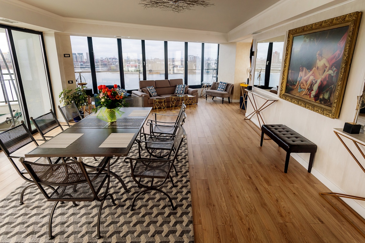 A spacious dining area is filled with natural light from large floor-to-ceiling windows. A modern wooden table is surrounded by six black chairs, while a colorful flower arrangement adds a touch of vibrancy. Elegant artwork adorns the walls, enhancing the room's modern design.