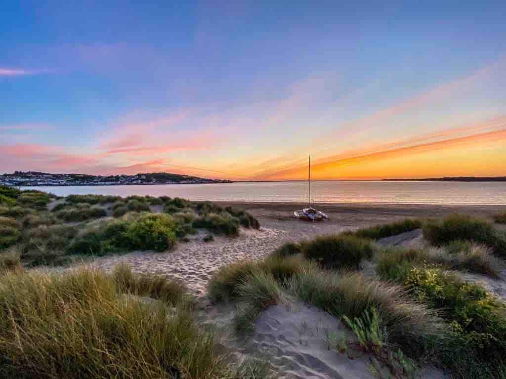 A tranquil beach scene features soft sand dunes dotted with lush, green grass under a vibrant sunset. The horizon showcases a gradient of warm colors, while a small sailboat rests peacefully on the water, completing the serene coastal view.