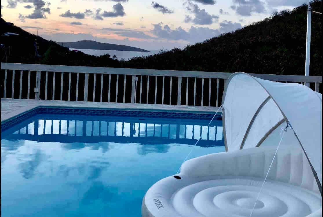 A large swimming pool reflects the evening sky, with gentle waves visible in the distance. A white float with a sunshade rests on the water's surface, inviting relaxation. Lush hills frame the backdrop, enhancing the serene atmosphere.