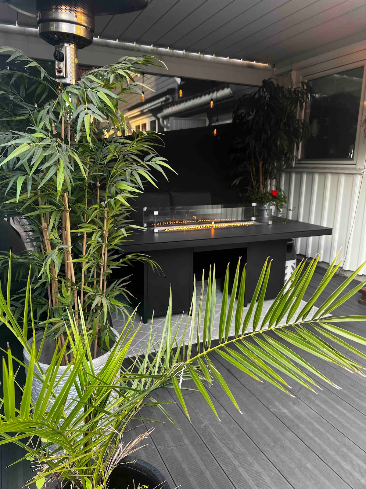 An outdoor area features a modern gas heater and a sleek tabletop surrounded by lush greenery. Tall bamboo plants and potted foliage create a serene atmosphere, while a wooden deck provides a spacious floor, ideal for gatherings.