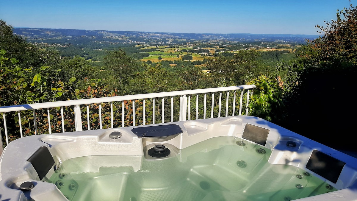 A jacuzzi is positioned on a terrace overlooking a scenic valley, offering expansive views of the surrounding landscape. The clear water reflects the sky, and the area is enveloped by greenery, enhancing the sense of tranquility and privacy.