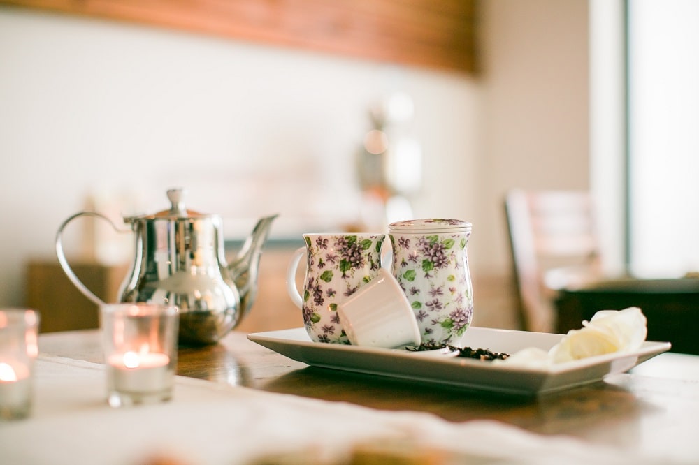A set of floral-patterned cups and a silver teapot are arranged on a table. Accompanying the cups is a rectangular plate with slices of banana. Small candles create a soft glow in the background, enhancing the serene setting.
