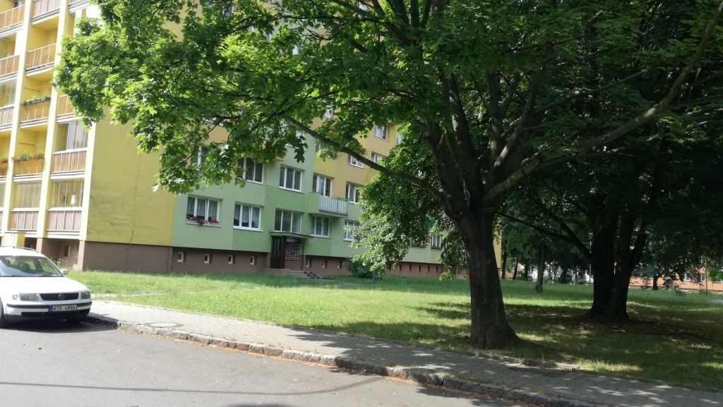 The exterior of a multi-story building is visible, featuring light green and yellow façades. Large windows enhance the modern design. Mature trees provide shade around a grassy area in front of the building, contributing to a serene environment.