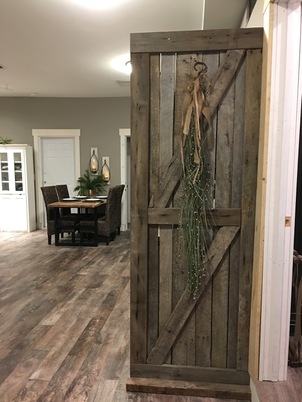 A rustic wooden sliding door is displayed, adorned with a natural greenery accent. In the background, a dining area is visible with a small table and woven chairs. Light wooden flooring adds warmth to the spacious gathering area.