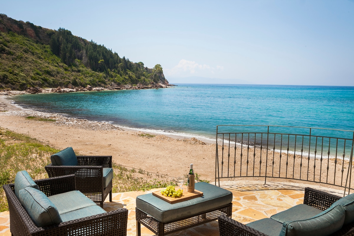 A patio area features comfortable seating arrangements facing the beach, with a low table displaying fruit and a drink. The clear blue waters of Lourdas Bay and sandy shore are visible, framed by lush greenery and distant hills under a bright sky.