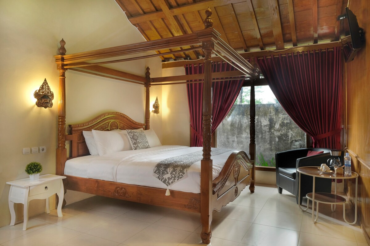 A traditional four-poster bed crafted from wood is featured in the sleeping area, accompanied by crisp white linens. Dark curtains frame the window, allowing natural light to filter in. A small side table and a black armchair are positioned near the window, enhancing the cozy ambiance.