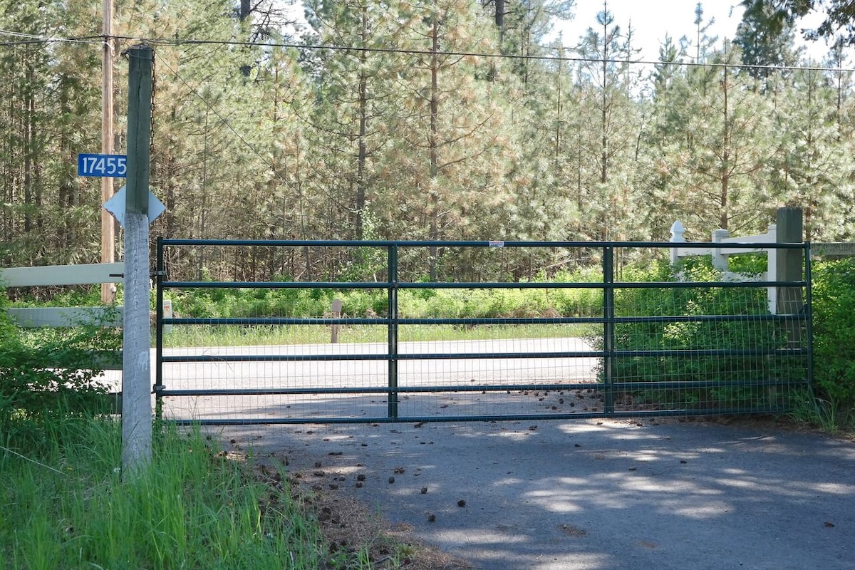 A green metal gate is positioned at the entrance of the property, surrounded by greenery. A blue address sign is attached to a nearby post. The road beyond the gate leads into a wooded area, with tall trees partially visible in the background.