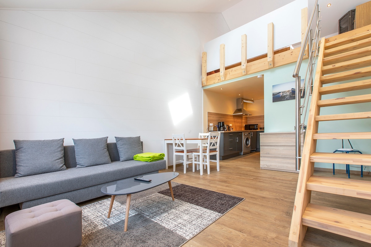A spacious living area features a gray sofa complemented by a green throw, a low wooden coffee table, and a soft area rug. A dining space with white chairs and a kitchen area is visible in the background, with wooden stairs leading to the mezzanine above.