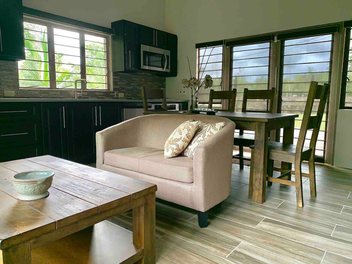 An open kitchen and dining area showcase modern amenities and natural light. A beige sofa with decorative pillows complements a wooden dining table surrounded by four chairs. Large windows provide views of the outdoors, enhancing the spacious feel of the room.