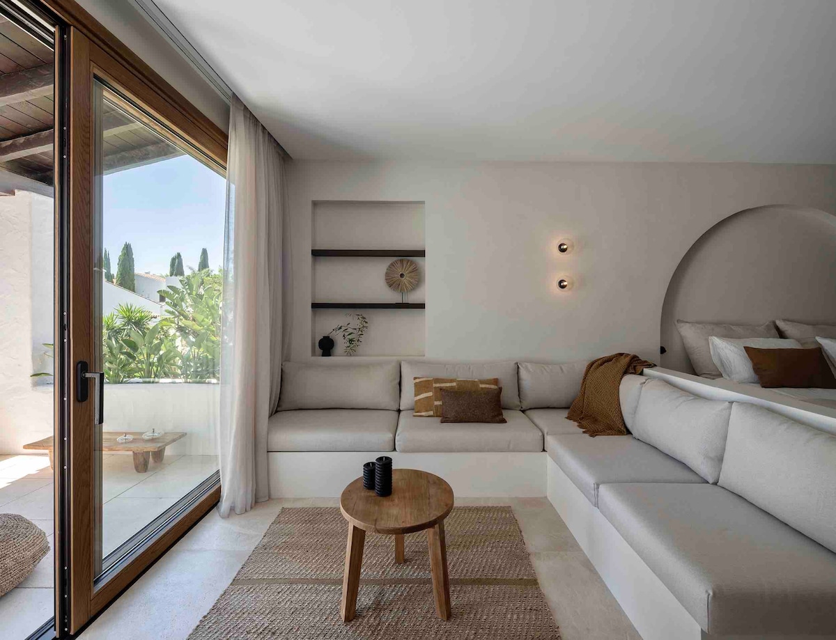 A modern living area is highlighted by a spacious, L-shaped sofa, complemented by a round wooden coffee table. Natural light filters through large glass doors, offering a view of the outdoor greenery. Minimalist decor is used to create an inviting atmosphere.
