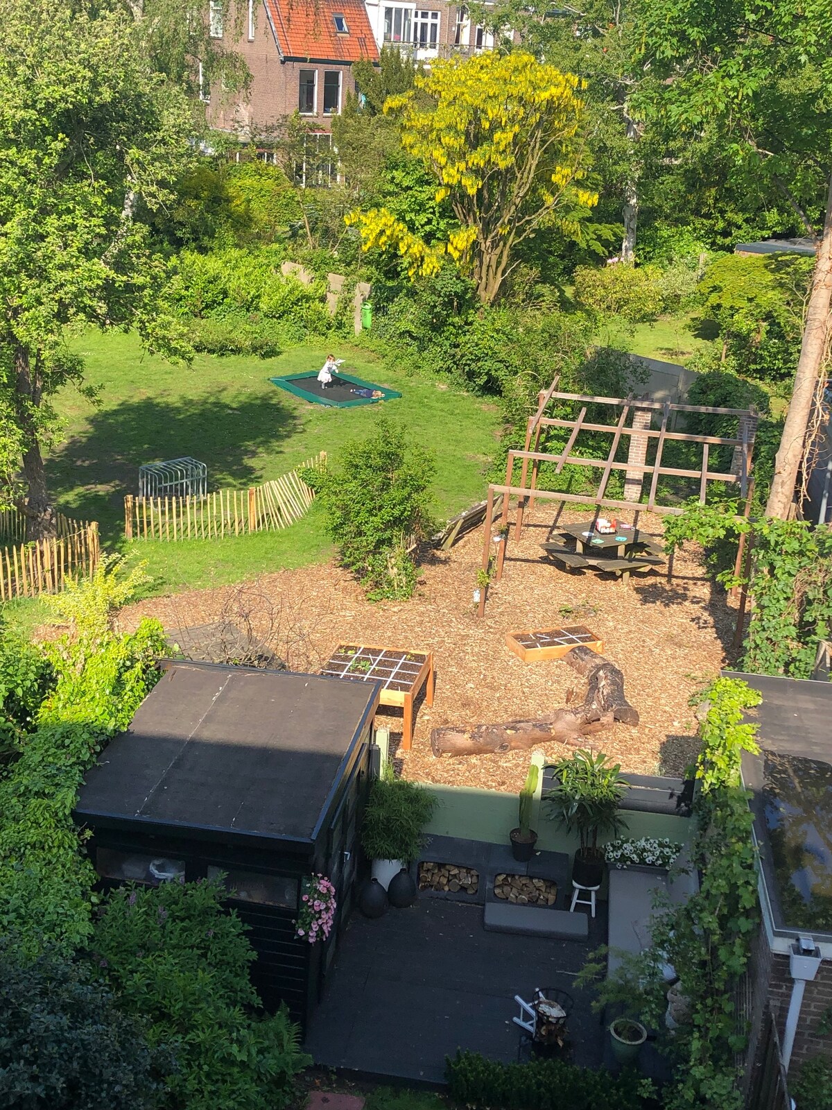 A spacious garden is depicted, featuring a wooden play structure and a sandbox area surrounded by greenery. A grassy area is seen in the background with a picnic blanket laid out. Various plants and a wooden storage shed are present, enhancing the outdoor space's charm.