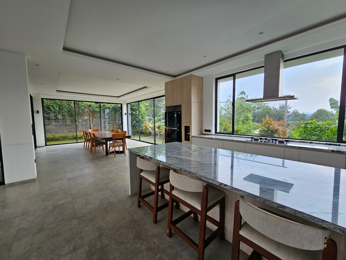 A modern kitchen features a large island with seating and a built-in cooktop. Expansive glass walls allow natural light to fill the space and provide views of the greenery outside. A dining area with a wooden table is visible in the background.