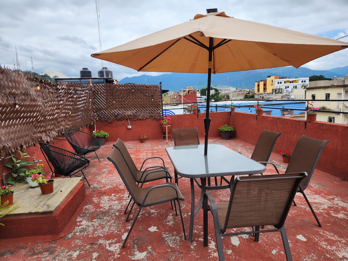A spacious terrace features a large umbrella providing shade over a dining table surrounded by several chairs. Colorful planters with plants are arranged around the space, with a backdrop of rooftops and mountains visible in the distance.