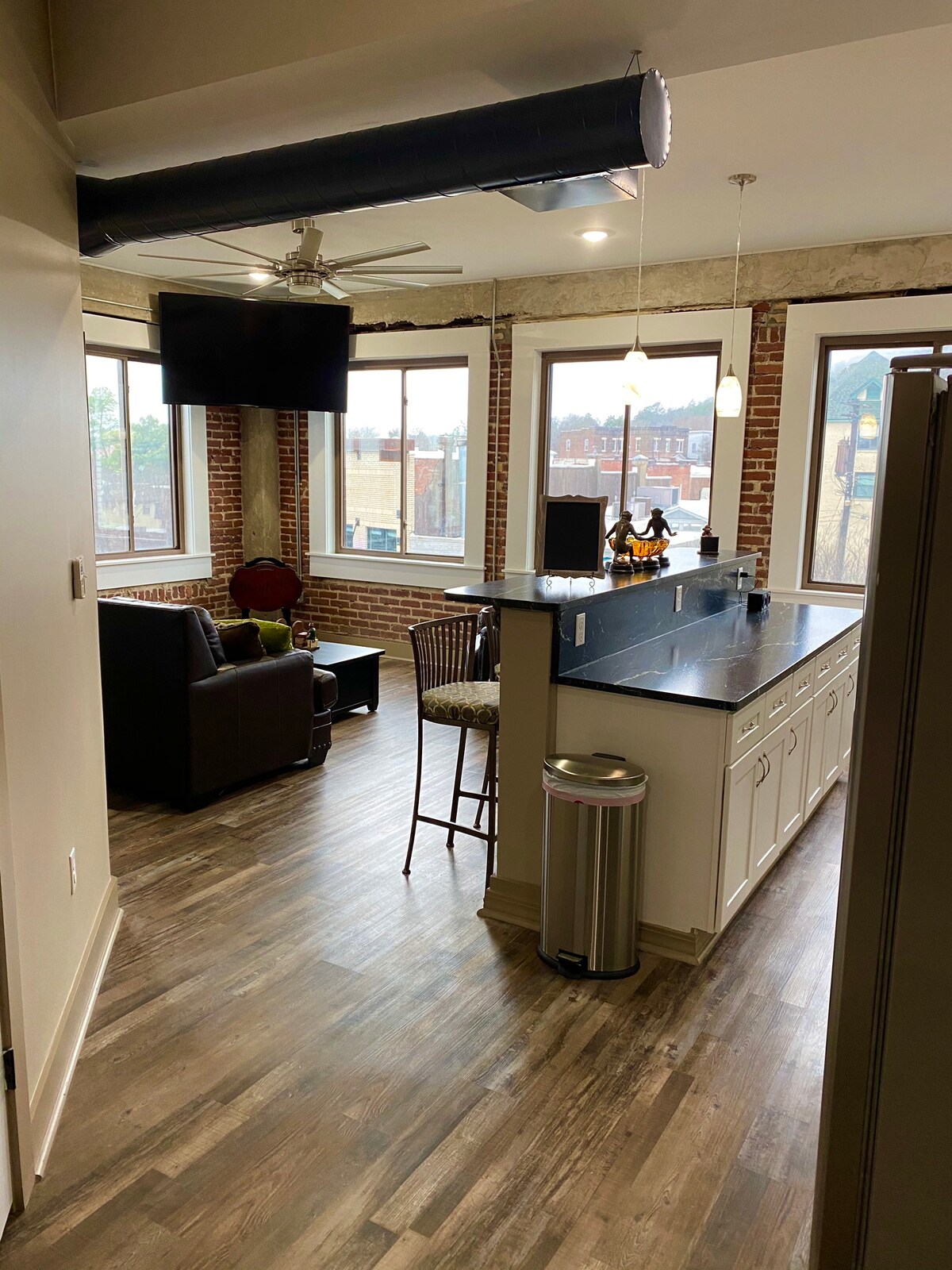An open-concept living area is shown with exposed brick walls and large windows, allowing natural light to fill the space. A bar counter with two stools is positioned near a modern kitchen, while a cozy seating area featuring a dark armchair and small table is visible.