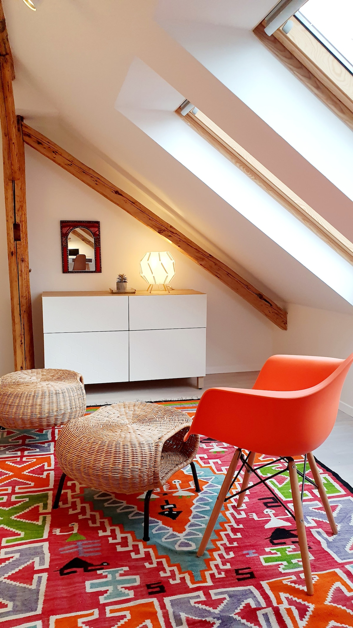 A stylish nook is highlighted with a vibrant area rug featuring geometric patterns. Two woven stools sit alongside an orange chair. Natural light floods the space through angled skylights, illuminating a modern white cabinet and a mirror mounted on the wall.