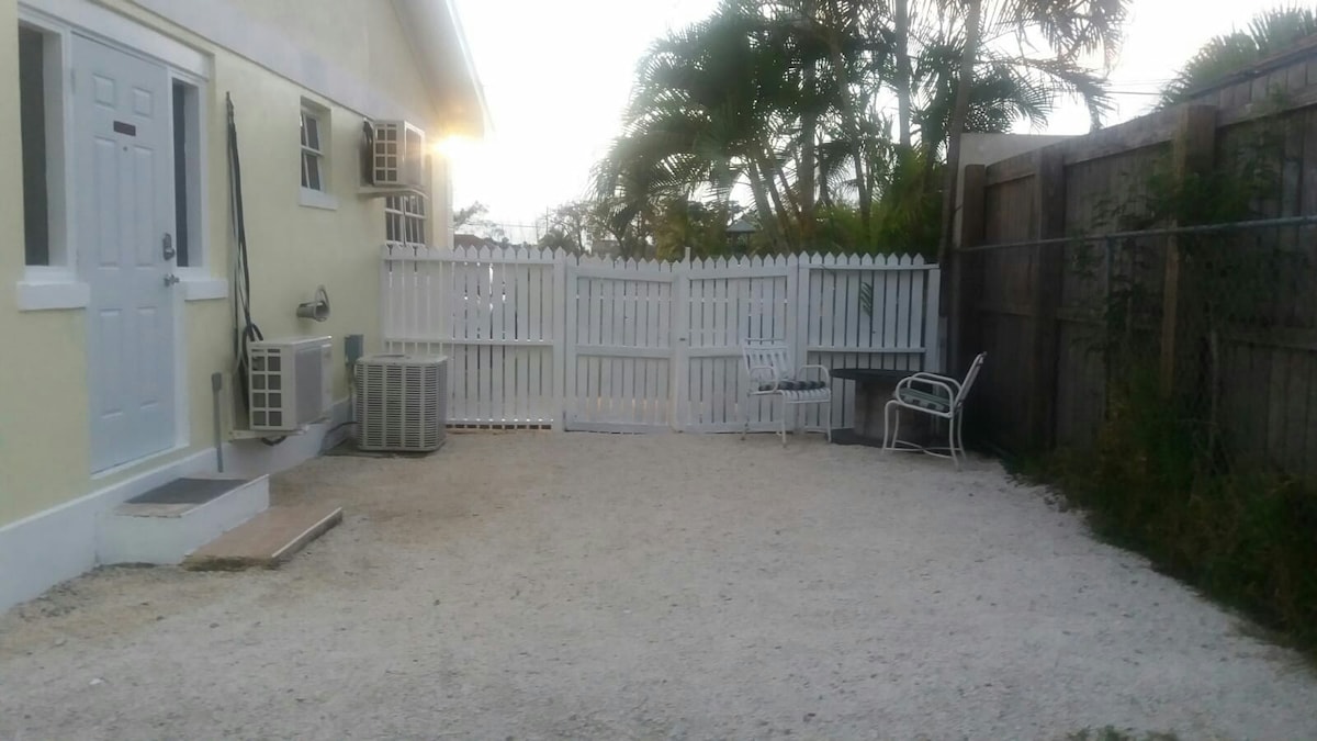 A private outdoor space is visible, featuring a gravel surface bordered by a white picket fence. An air conditioning unit is positioned next to the entrance, while a small seating area with a white chair is located on the side, creating an inviting space for relaxation.