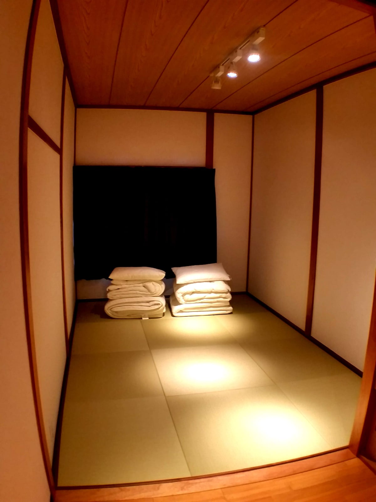 A minimalistic room features tatami flooring and wooden accents. Neatly stacked futons are placed against a wall, illuminated by soft lighting. A window covered with dark curtains is situated nearby, contributing to a serene and simple atmosphere.
