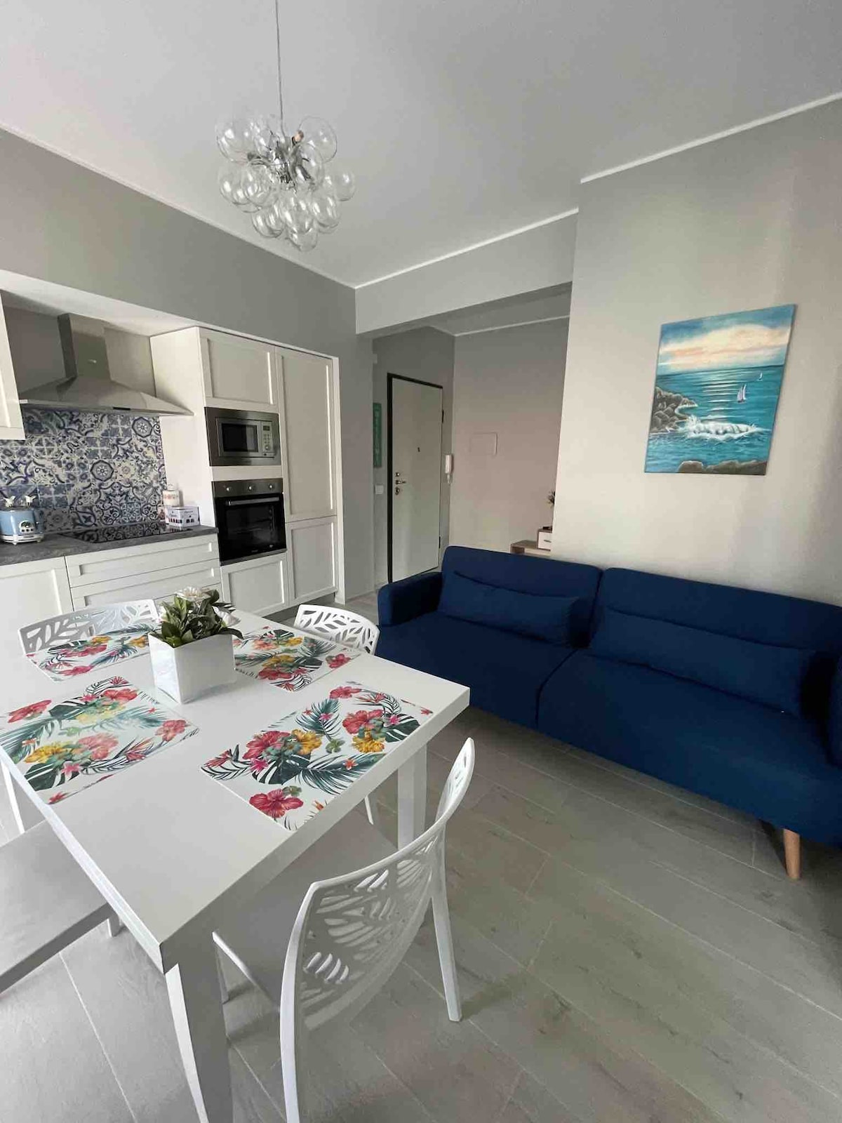 A modern dining area is seen with a white table set for four, featuring vibrant tropical-themed placemats. A navy blue sofa is positioned against the wall, complemented by a light-colored artwork depicting a coastal scene. A well-equipped kitchen area can be partially observed in the background.