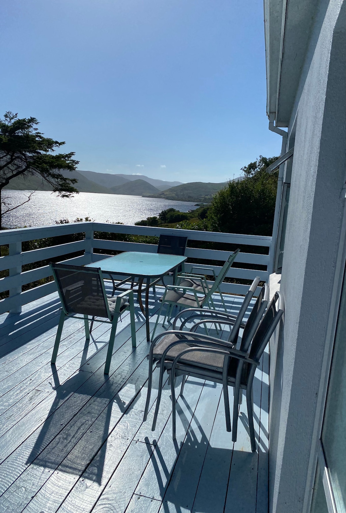 A balcony area features a light blue table surrounded by six chairs, offering a scenic view of the shimmering water and surrounding hills. The deck is illuminated by natural sunlight, enhancing the pleasant atmosphere of the outdoor space.